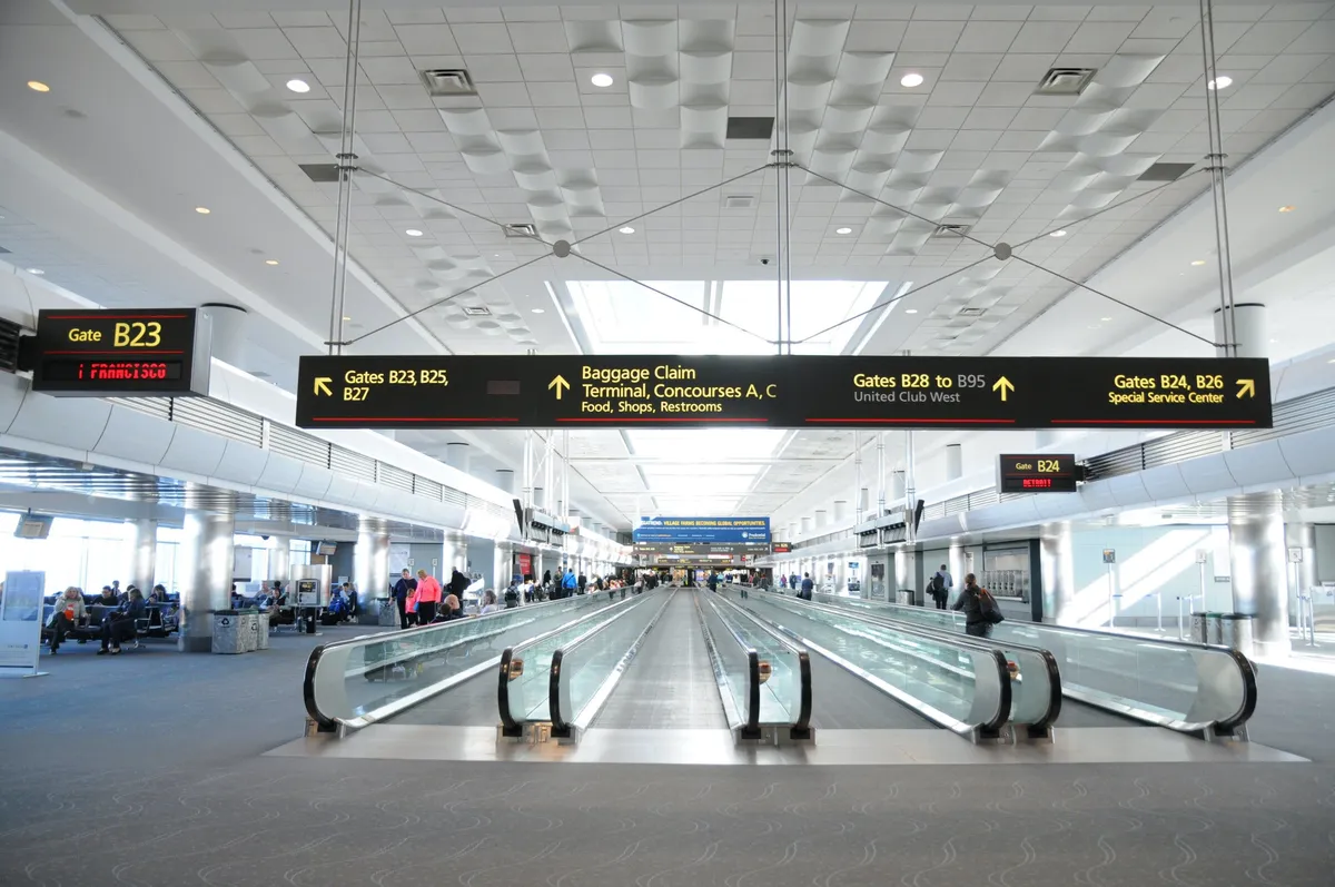 The image captures a bustling airport interior with moving walkways, directional gate signs, and open seating areas. Travelers move purposefully through the terminal, symbolizing the flow of visitors to and from business destinations. The mood is energetic and anticipatory, echoing the convenience and connectivity essential for business guests.