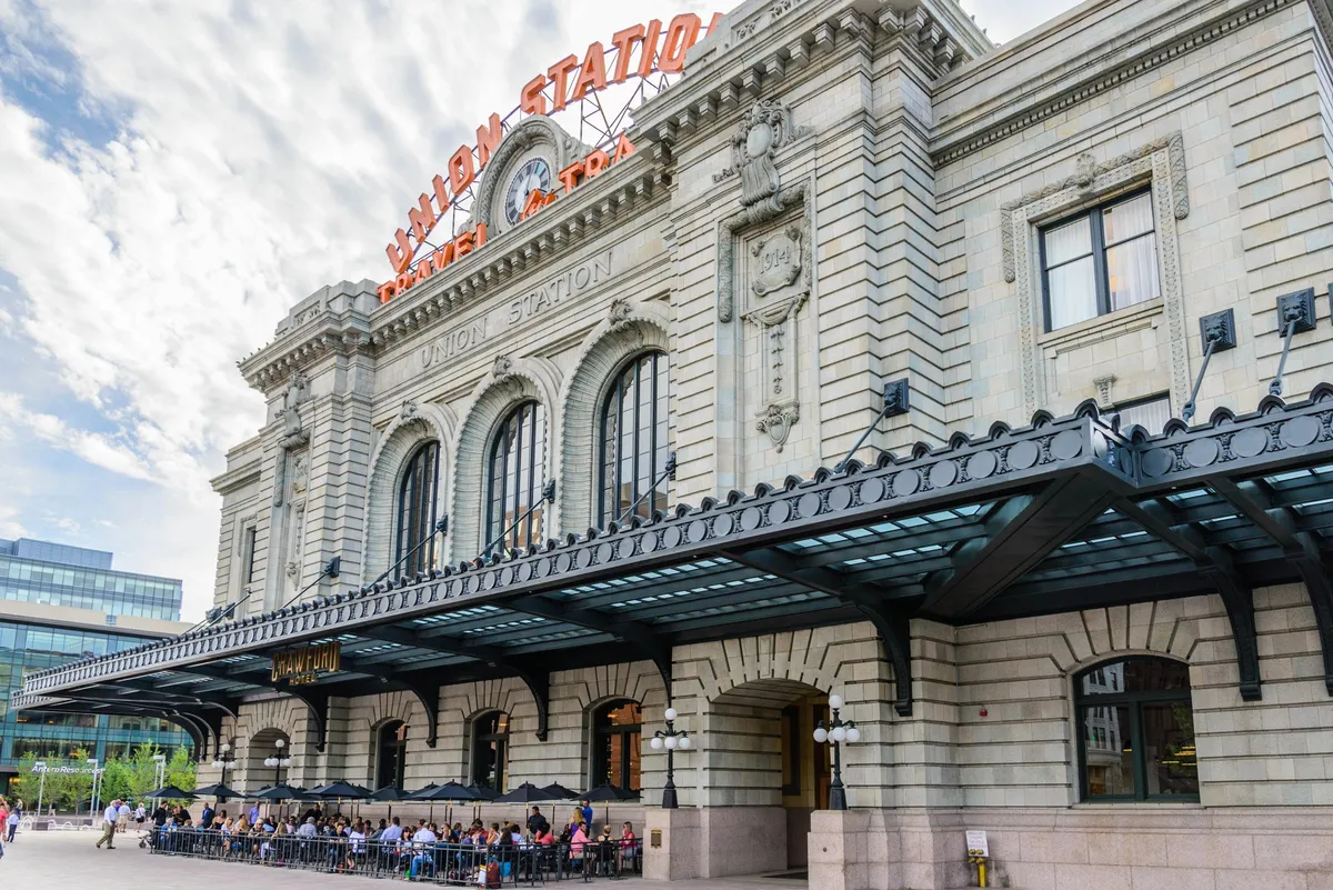 A vibrant street scene featuring the grand façade of Denver's historic Union Station, illuminated by the prominent 'UNION STATION' sign. People are seen enjoying meals in the lively outdoor seating area, adding a welcoming, urban vibe.