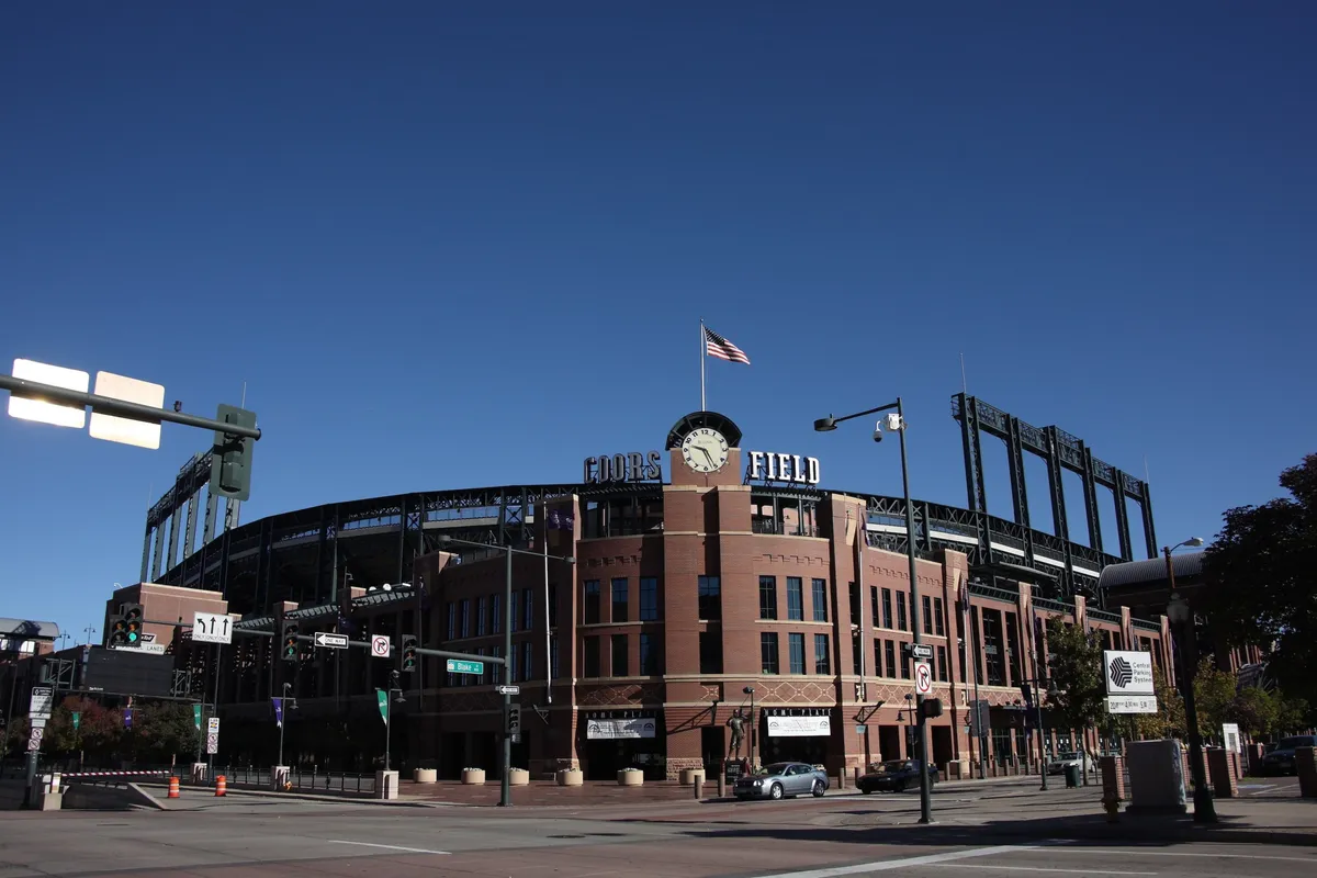 The image shows the exterior of Coors Field, with a prominent Coors Field sign, a large clock on the building, and an American flag waving above the stadium structure. The area buzzes with energy, capturing a sense of excitement and vibrancy as people gather for events or explore the lively neighborhood surrounding the venue.