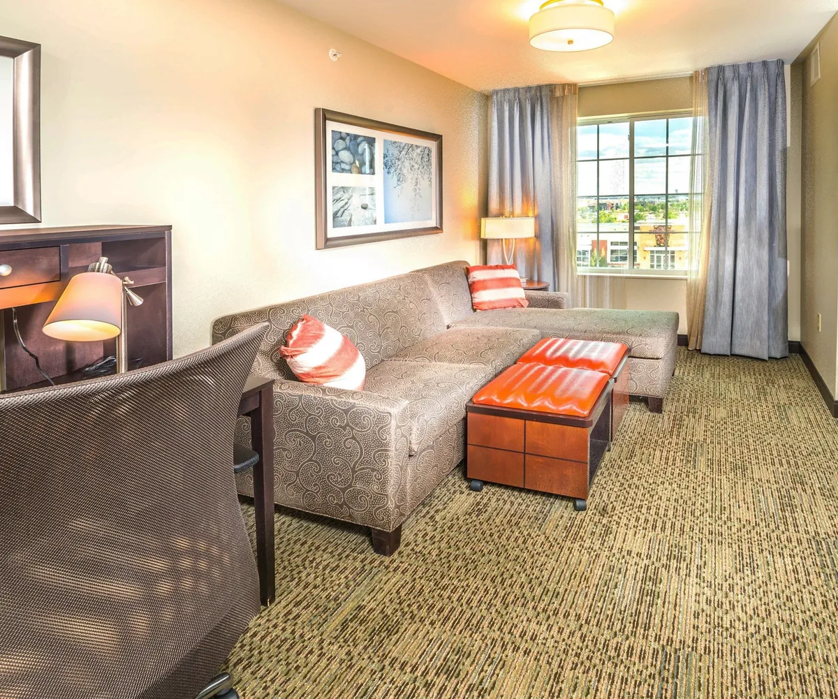 A warmly lit living area with a comfortable sofa, stylish coffee table, and a dedicated work desk, all bathed in natural sunlight from a large window. The space feels cozy and inviting, ideal for relaxing, socializing, or catching up on work during your Denver getaway.