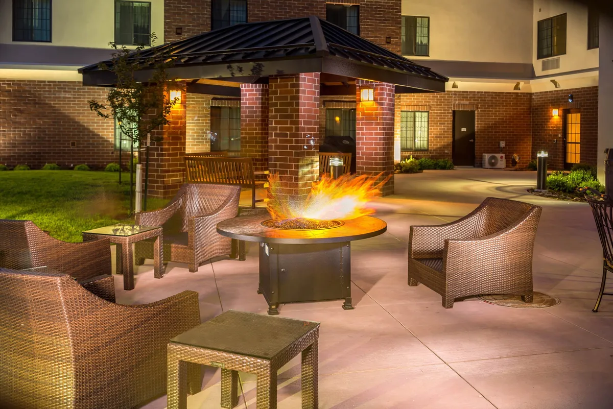 An inviting outdoor BBQ area with a fire pit surrounded by comfortable seating, accent lighting, and lush landscaping. The setting suggests a social, relaxed environment ideal for grilling, gathering, and sharing stories under the open sky.