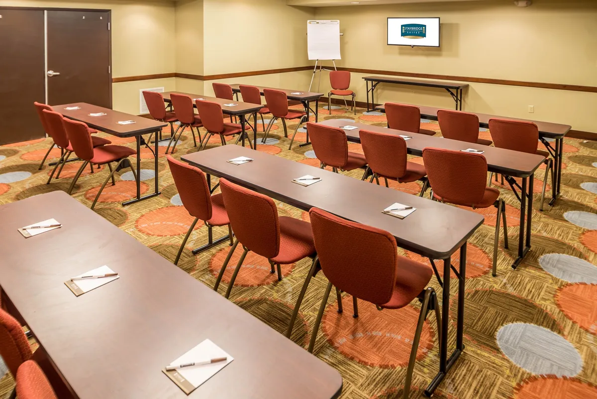 The image captures a bright, well-organized interior space with rows of tables and chairs arranged to facilitate group gatherings or collaborative work. A presentation screen and whiteboard are visible at the front, emphasizing the room's suitability for meetings, workshops, or team sessions. The overall mood is focused and welcoming, with ample lighting and a layout that encourages interaction.