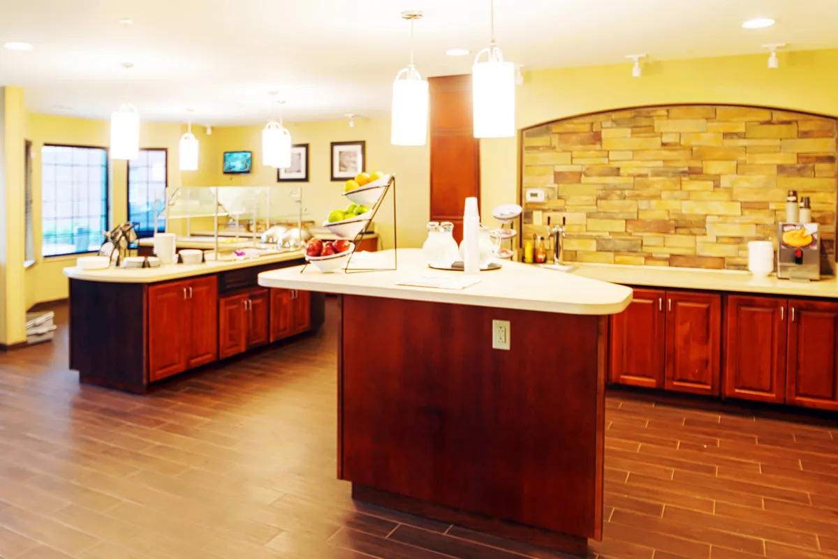 An inviting open-plan dining and living area with a central island, fruit display, and a cozy seating arrangement. The stone wall accent and warm lighting create a friendly, homey atmosphere ideal for gathering, sharing meals, or relaxing over coffee.