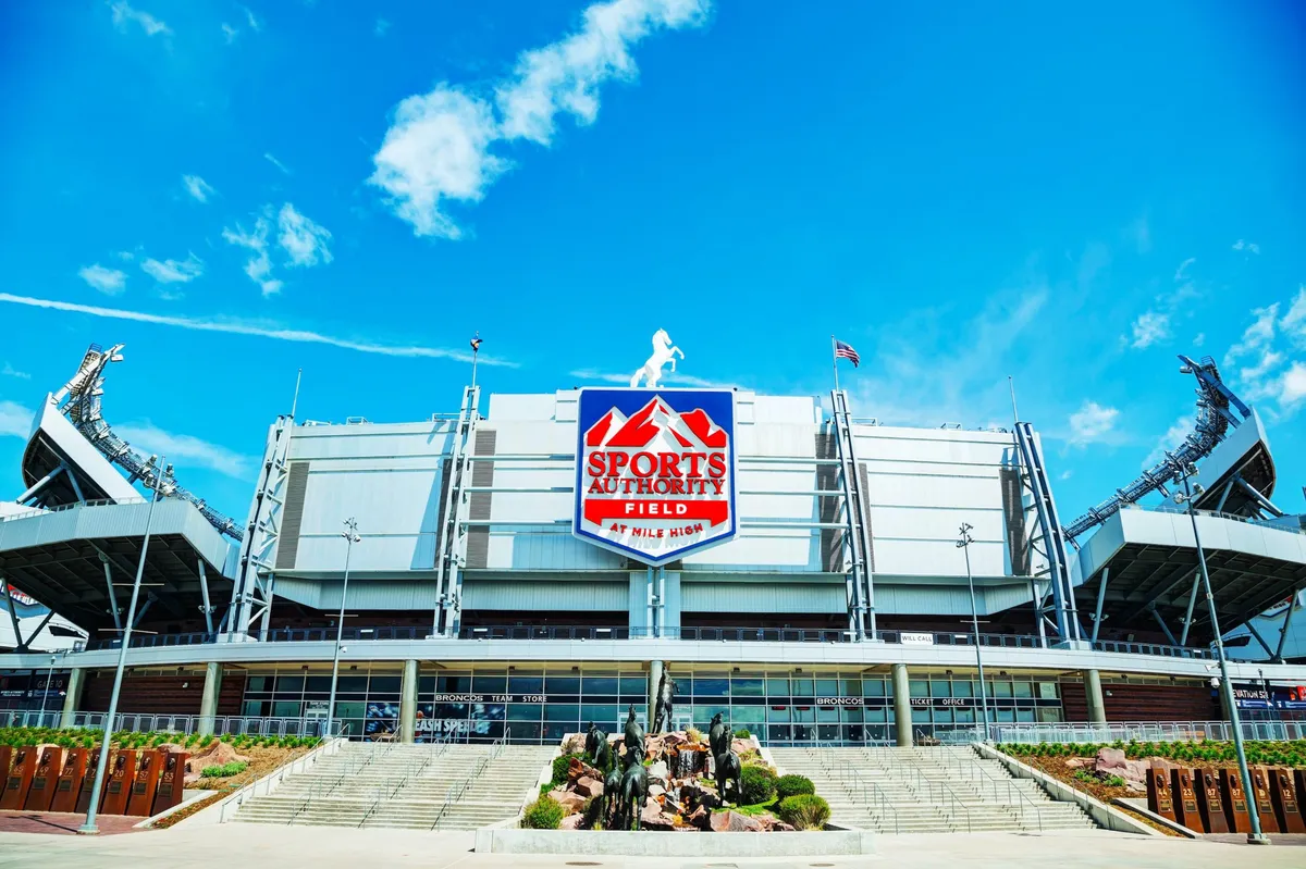 The image captures the impressive exterior of Sports Authority Field, with its recognizable sign, the Rocky Mountain statue at the forefront, and the dramatic stadium architecture. It exudes the anticipation and excitement associated with attending large-scale sports events or concerts.