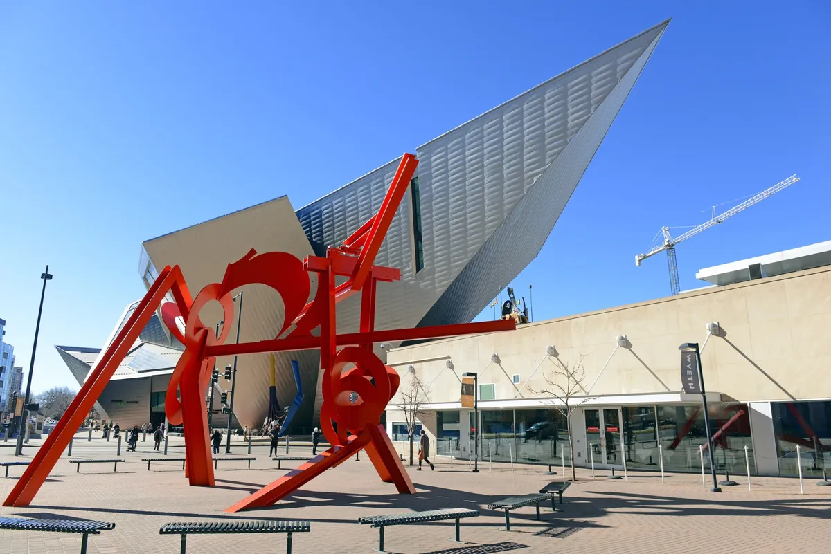 A modern Denver streetscape with a large, striking red sculpture in front of a contemporary building. Families and individuals can be seen strolling and interacting with the public art, suggesting a lively and culturally rich environment.